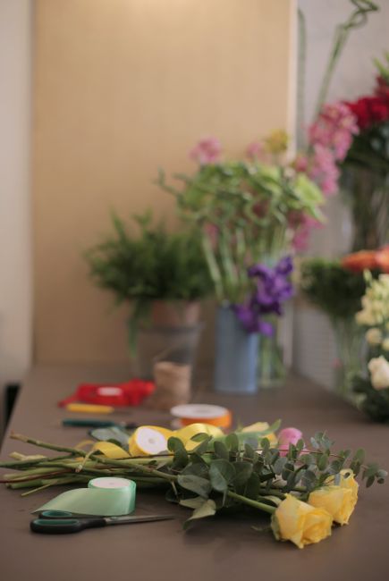 A close-up view of a florist's workspace featuring a partially assembled bouquet of fresh yellow roses and green foliage, with floral scissors, floral tape, and wrapping materials such as paper and ri