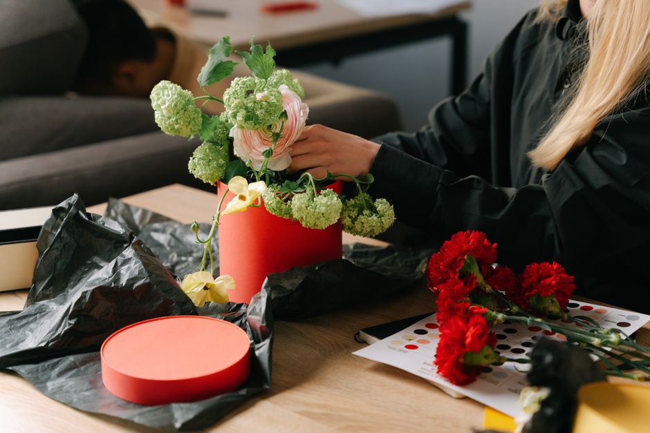 A person arranging a flower bouquet with a mix of pale pink roses, green hydrangeas, and trailing yellow orchids in a coral cylindrical vase on a wooden table. The flowers appear fresh with vibrant pe