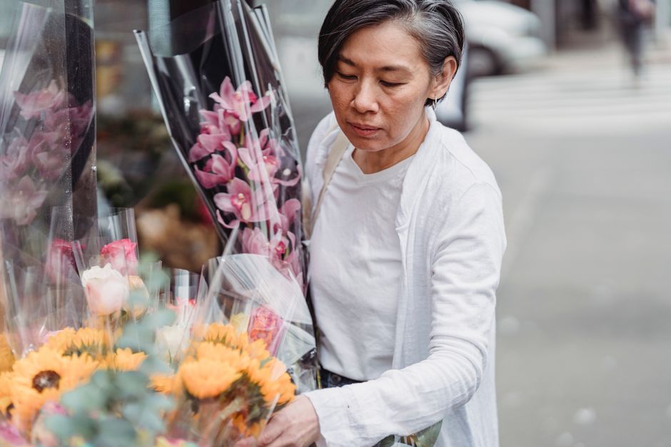 An older woman with short gray hair, dressed in a white shirt and light-colored cardigan, stands outdoors on a city street examining a bouquet of fresh flowers wrapped in clear plastic. The bouquet fe