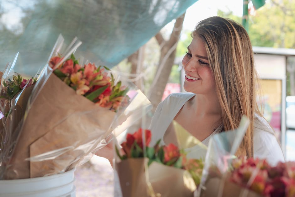 A young woman with long, light brown hair and a bright smile stands outdoors near a tree, selecting a fresh flower bouquet from a display of floral arrangements wrapped in brown paper and clear plasti