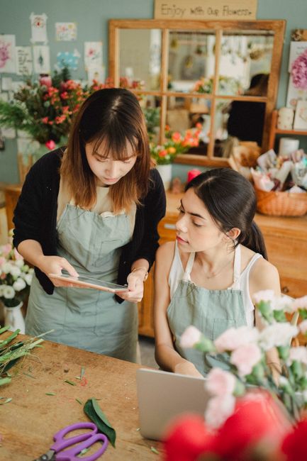Two women in a florist workshop at flowerscubitttown.co.uk discussing floral arrangements. One woman with shoulder-length brown hair, wearing a black cardigan and light grey apron, holds a clipboard a
