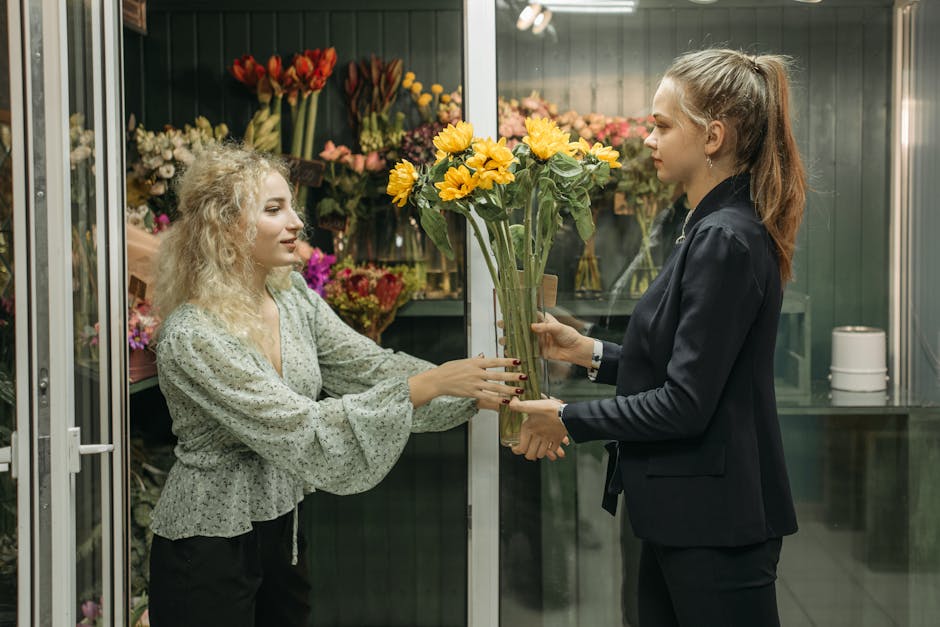 A florist with shoulder-length blond hair, wearing a black blazer and black trousers, is handing a tall bouquet of vibrant yellow sunflowers and mixed green foliage to a customer with curly blonde hai