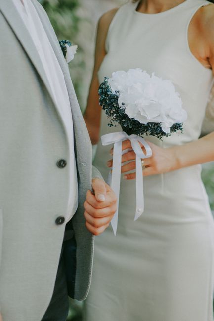 A bride holding a bouquet of white hydrangeas and dark greenery, tied with a white satin ribbon, standing next to a groom dressed in a light grey suit with a white shirt and grey tie. The bouquet is p
