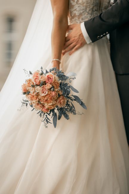 A close-up of a bride and groom during a wedding ceremony, with the bride holding a floral bouquet featuring blush pink roses, peach carnations, and blue-grey eucalyptus leaves. The bouquet is wrapped