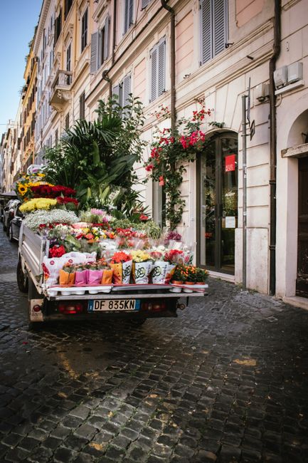 A delivery truck parked on a cobblestone street in an urban area, loaded with an array of fresh flower bouquets and potted plants. The arrangement includes vibrant roses, sunflowers, lilies, and green