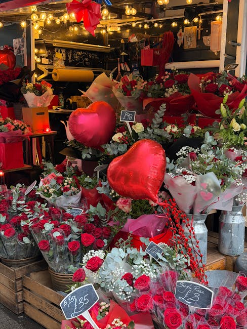 A vibrant display of floral gifts featuring red roses, white roses, and greenery arranged in bouquets and wrapped in pink and white tissue paper, with some in small metal buckets. Two large red foil h